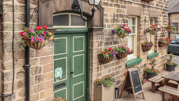 A stone building with a green door, hanging flower baskets, and a chalkboard sign. - Milford Cottage (Belper)