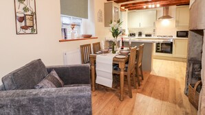 A cozy living room with a grey sofa, wooden dining table, and chairs. A kitchen with wooden beams and modern appliances is visible in the background. - Milford Cottage (Belper)