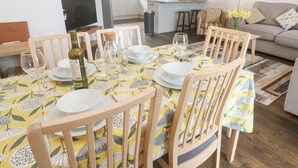 A dining area with a table set for four, surrounded by wooden chairs. A kitchen is visible in the background with white cabinets and a countertop. - Ocean Breeze (Marazion)