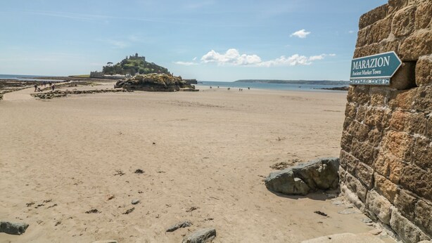 A beach with a sign reading "MARAZION Ancient Market Town" and a castle on a hill in the background. - Ocean Breeze (Marazion)