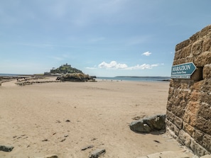 Ein Strand mit einem Schild, auf dem 'MARAZION Antike Marktgemeinde' steht, und einer Burg auf einem Hügel im Hintergrund.