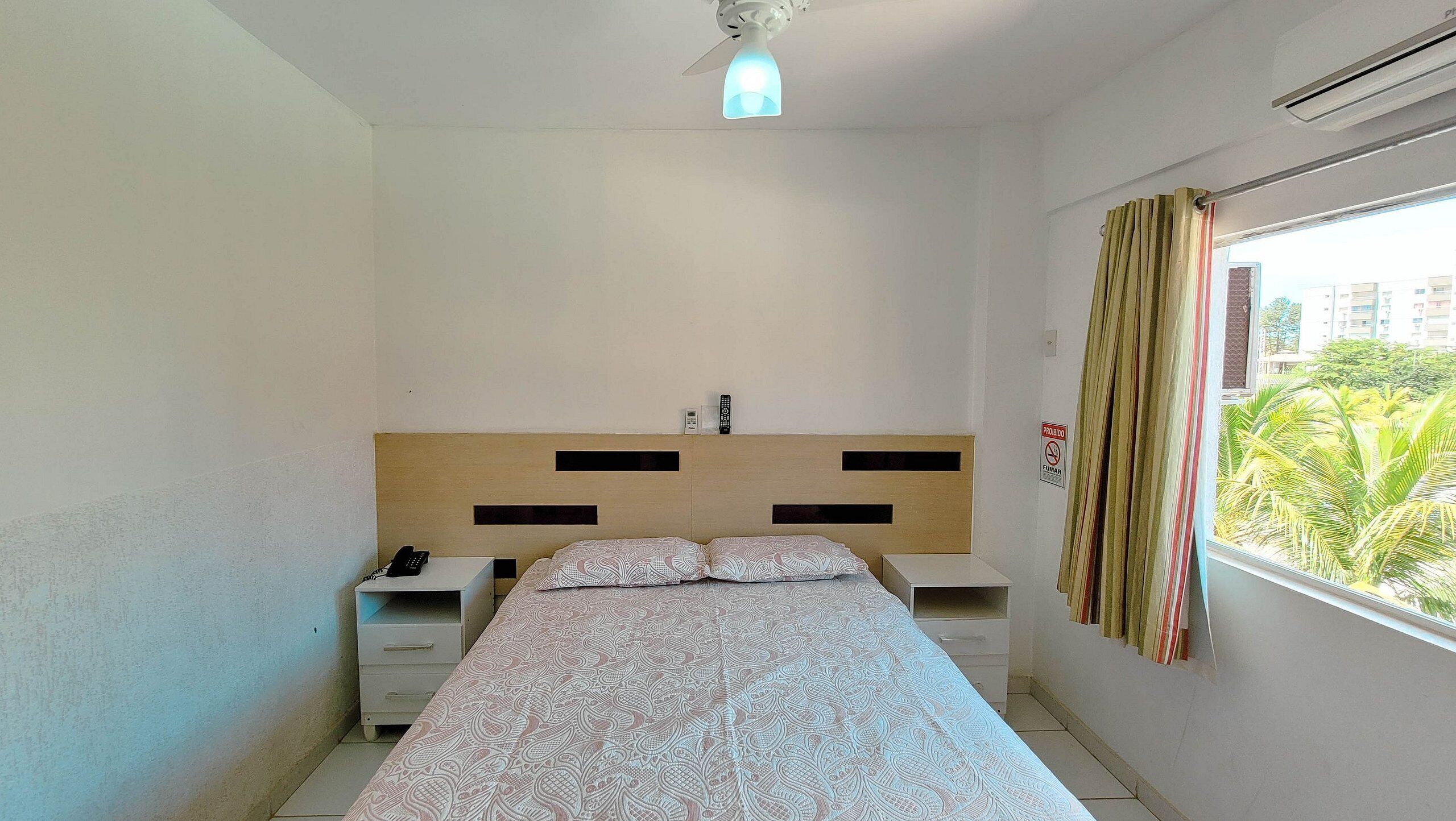 Un dormitorio con cama, mesitas de noche, ventilador de techo, ventana con cortinas y vista a zonas verdes.