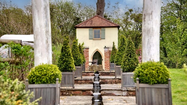 A small building with a tiled roof and a window with shutters, surrounded by a stone pathway and wooden fences. - The Folly (Hereford)