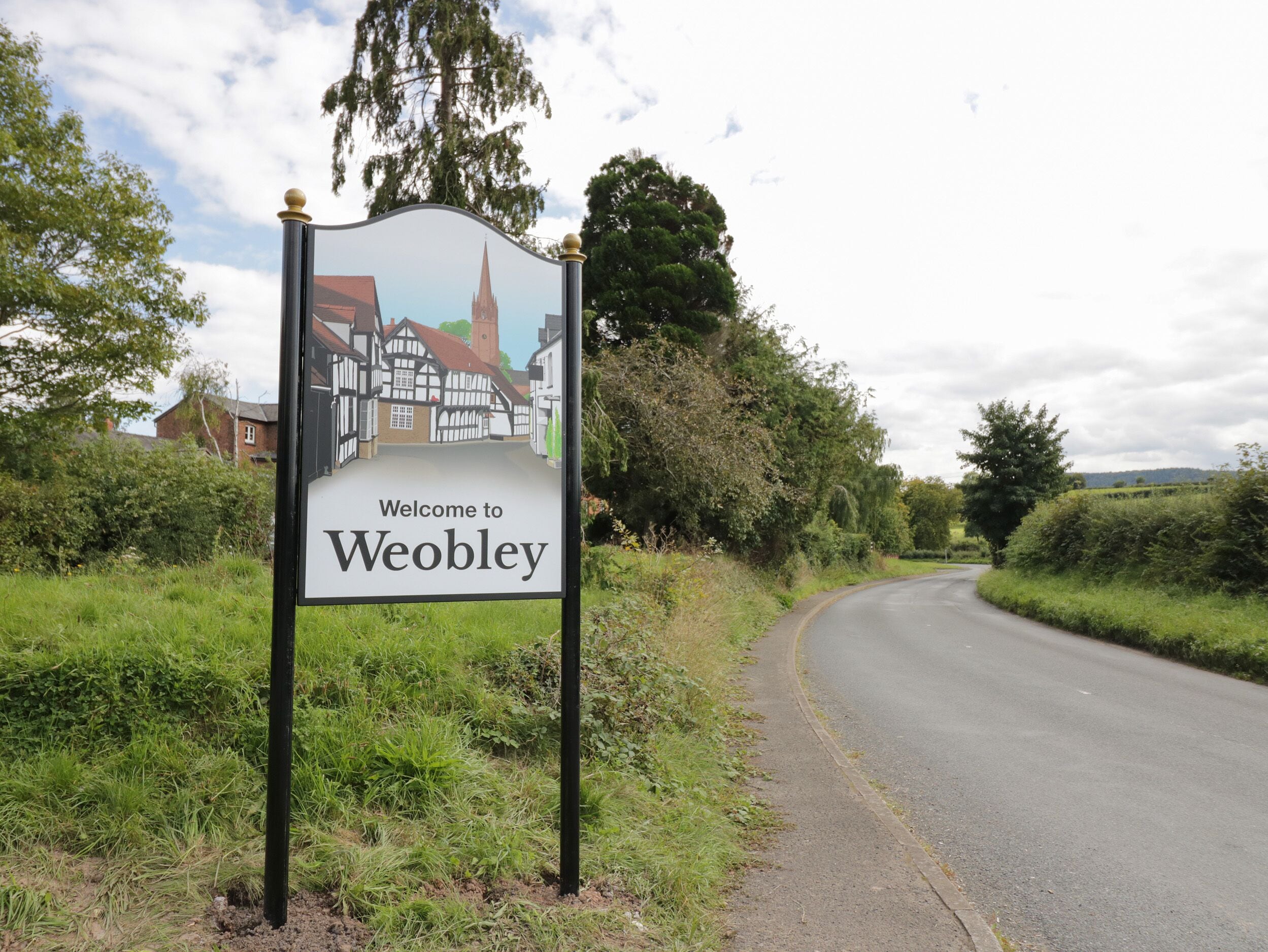 Un cartel de bienvenida a Weobley con una representación de edificios tradicionales y una iglesia.