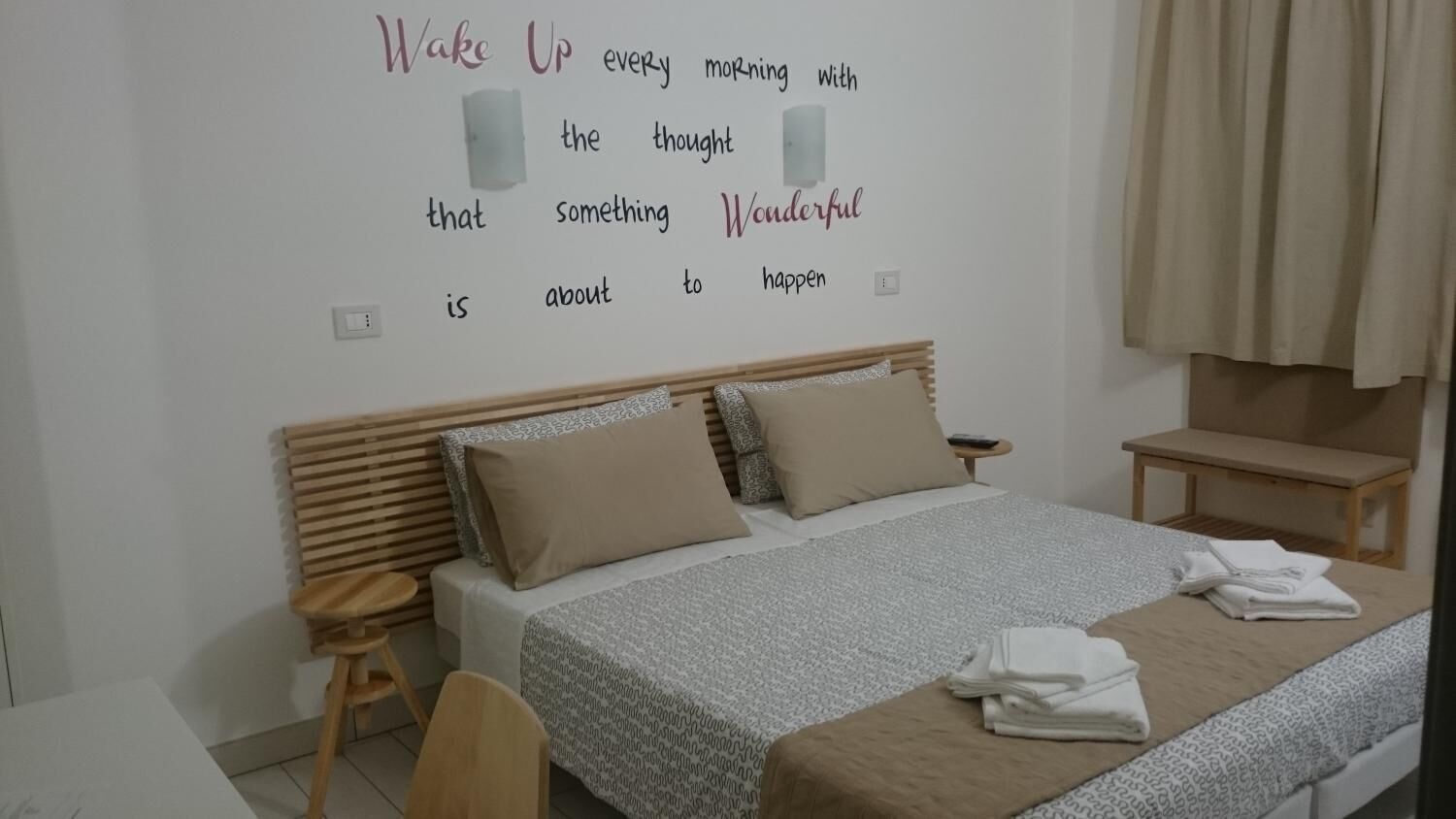 Un dormitorio ordenado con cama, almohadas, una mesita de noche y una pared con texto motivacional.