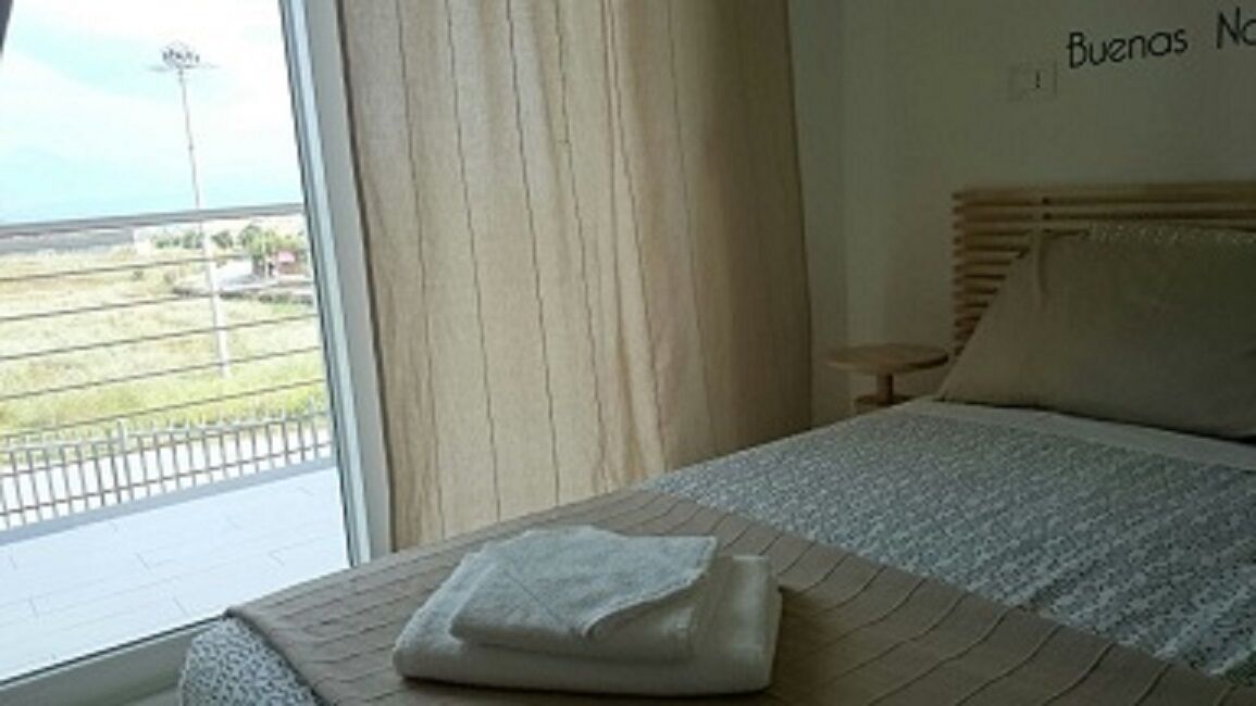 A hotel room with a bed, a window with curtains, a small table, and a wall sign.
