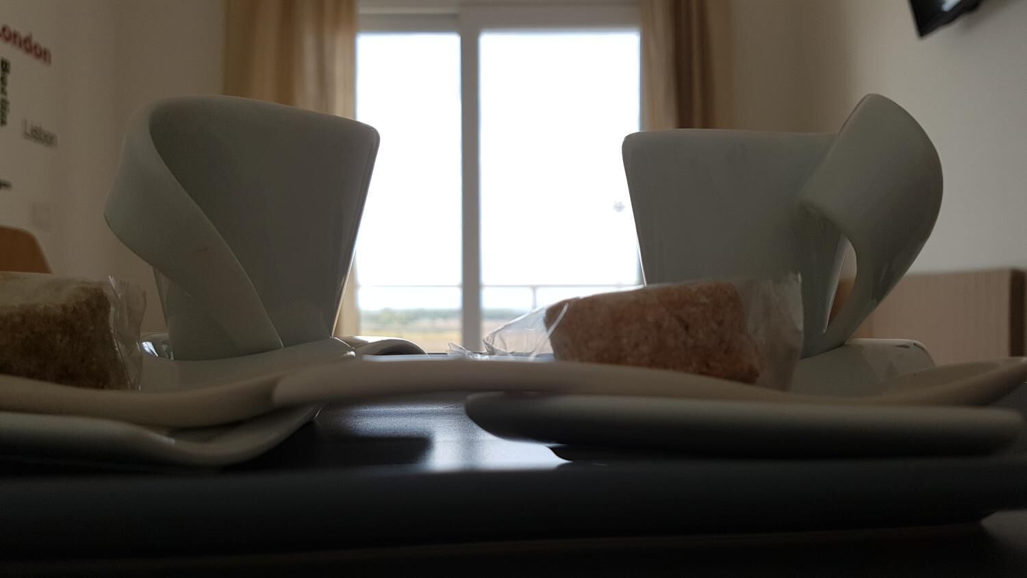 A hotel room with a view of the outdoors through a window, a bed with a white headboard, and a table with a cake and a glass.