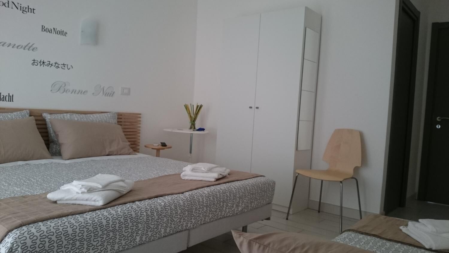A modern hotel room with a large bed, a chair, a small table, and a wardrobe.