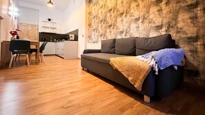 A modern living room with a sofa, wooden flooring, a kitchen in the background, and pendant lighting. - 3D FLORIANSKA 15 Apartament (Kraków)