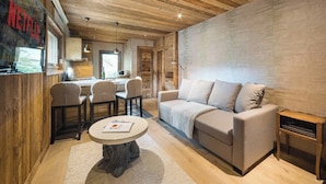 A cozy living room with a sofa, chairs, a coffee table, and a kitchen area visible through an open door. - Steps from Megève Rink & Chamois Gondolas (Megève)