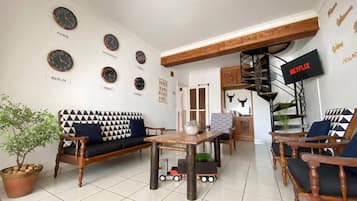 Una sala de estar con un sofá, sillas, una mesa de centro y un televisor. Hay relojes en la pared y una planta en el suelo.