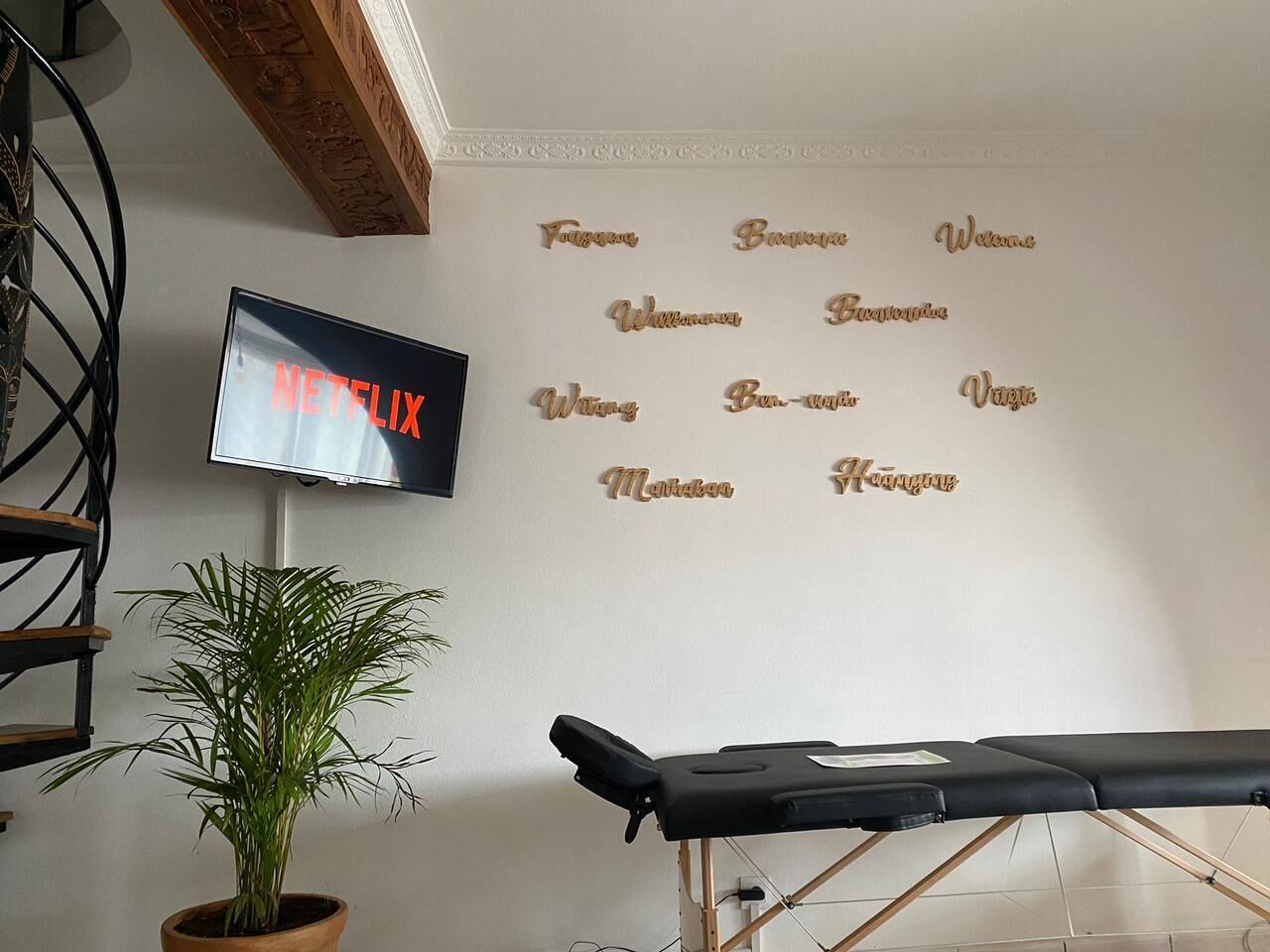 A room with a mounted TV displaying 'NETFLIX', a wall-mounted massage table, a potted plant, and a decorative wall with names.
