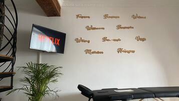 A room with a mounted TV displaying 'NETFLIX', a wall-mounted massage table, a potted plant, and a decorative wall with names.