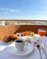 Mesa com itens de café da manhã, incluindo doces, frutas e um copo de suco, com vista para a cidade ao fundo.