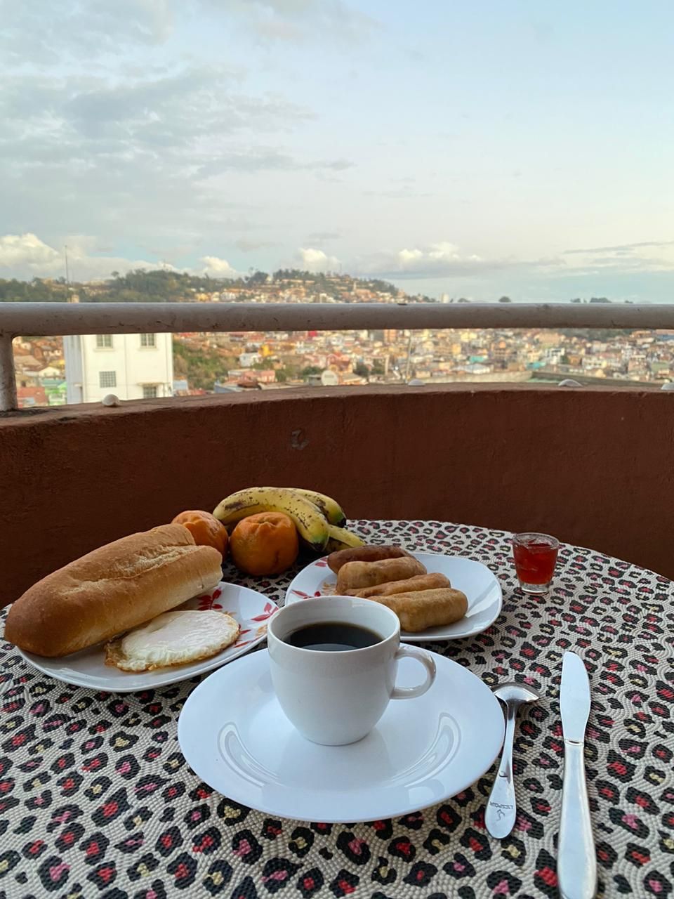 Una mesa con una taza de café, un plato de pasteles y un plátano en un balcón con vistas a un paisaje urbano.