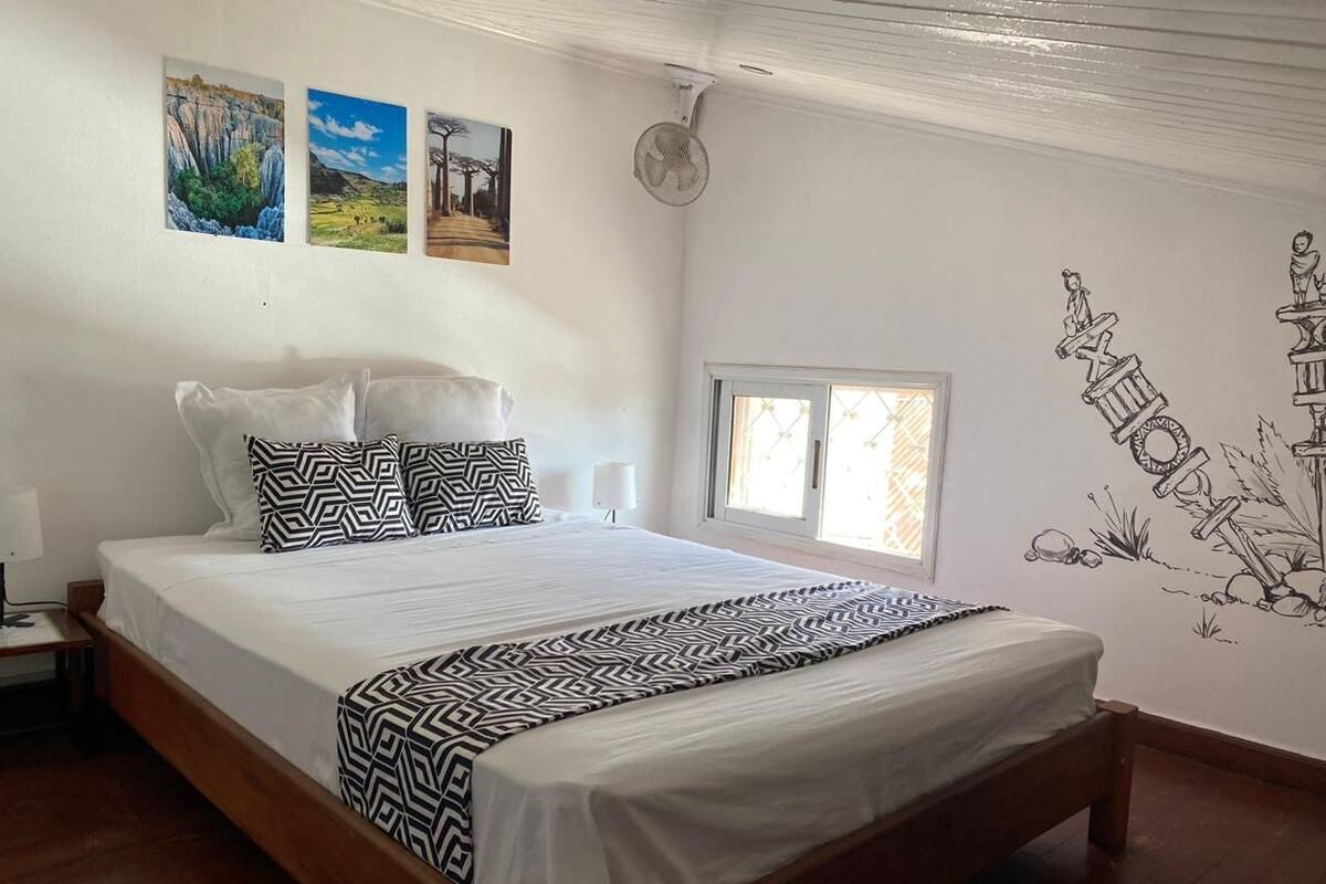 A bedroom with a wooden bed, white bedding, patterned pillows, a wall-mounted fan, and three landscape paintings.