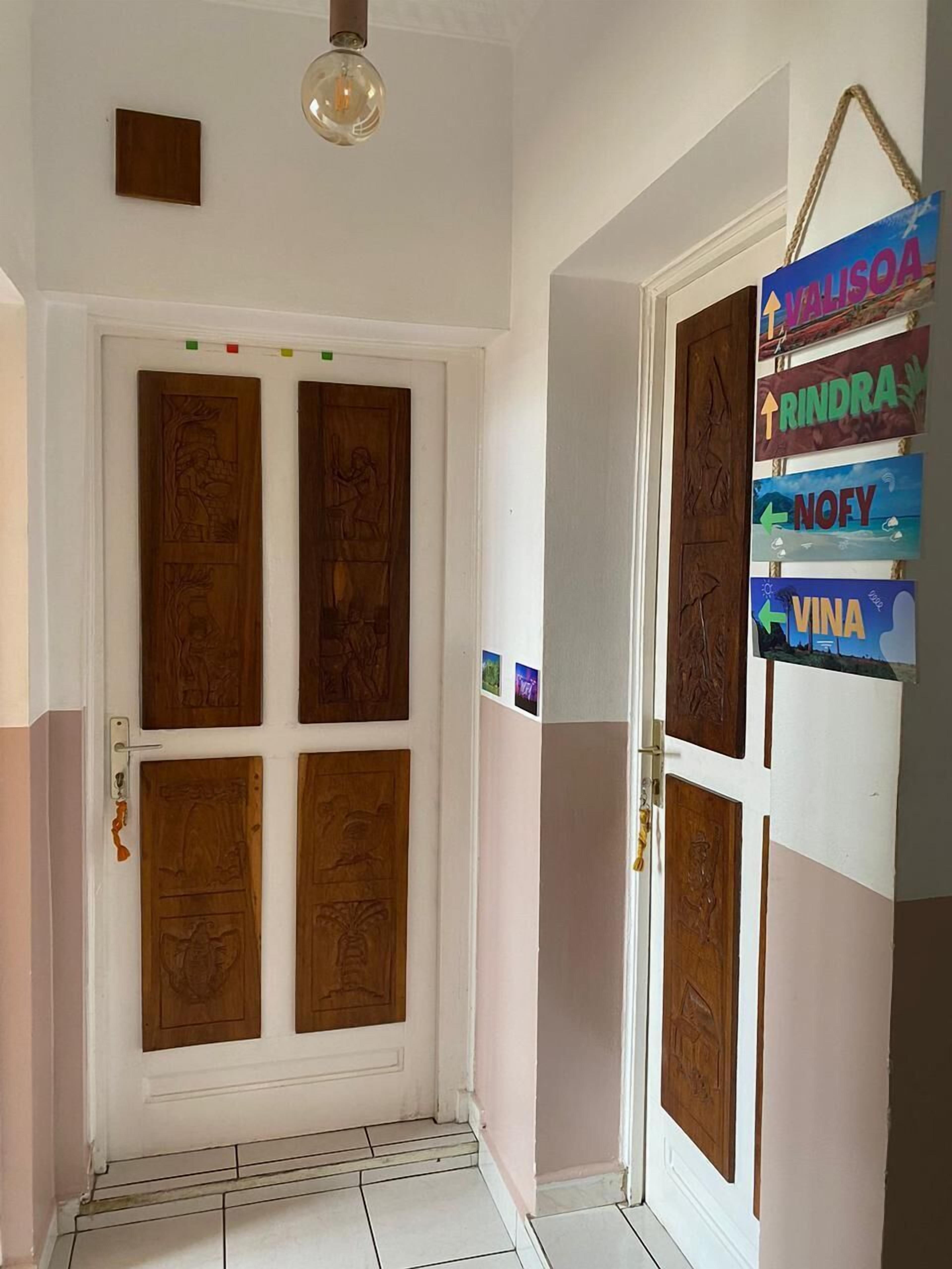 A hallway with a white door featuring four panels with intricate designs. A hanging sign with names and arrows is visible on the wall.