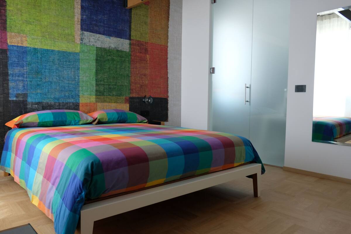 A bedroom with a colorful quilted bedspread, a wooden floor, and a large mirror on the wall.