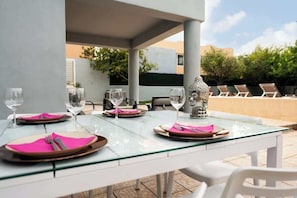 Un juego de mesa con cubierta de vidrio, adornado con servilletas rosas, cubiertos y copas de vino, con un jarrón decorativo y sillones de descanso al fondo.