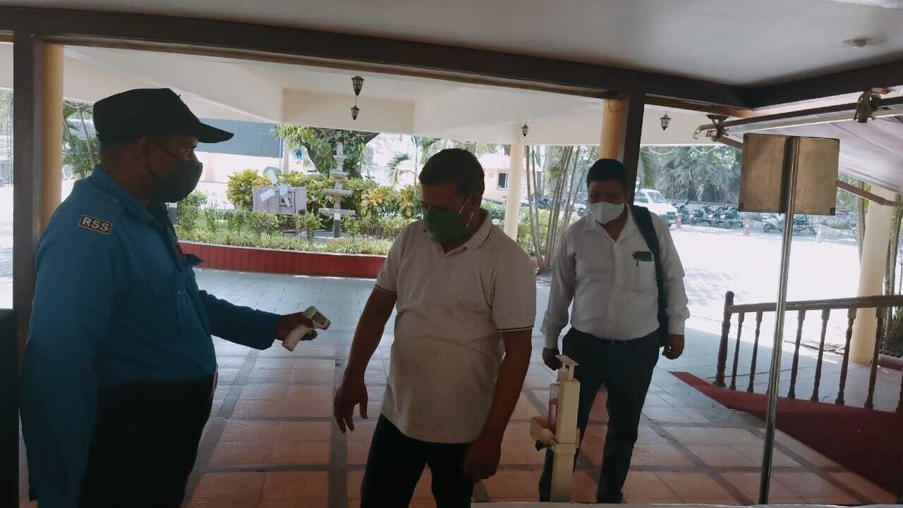 A man in a blue uniform standing and talking to two men in white shirts in a covered outdoor area with a tiled floor and a railing.