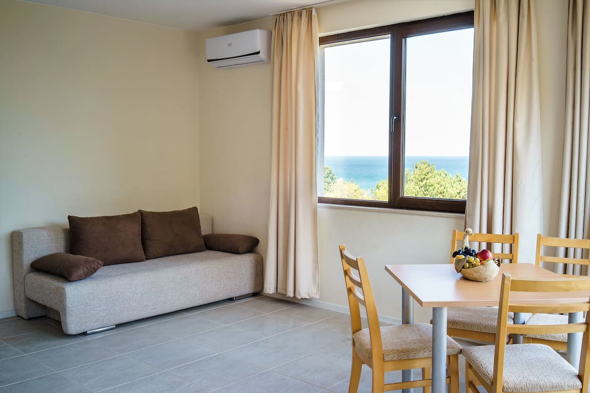 Ein Zimmer mit einer Couch, einem Esstisch mit Stühlen, einem Fenster mit Vorhängen und einer Klimaanlage.