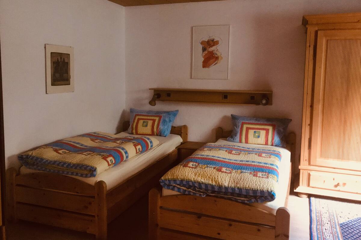 Quarto com duas camas de madeira, cada uma com manta e almofadas estampadas. Há uma cabeceira de madeira, um quadro emoldurado na parede e um armário de madeira.