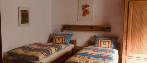 Une pièce avec deux lits en bois, chacun recouvert d’une couverture à motifs et agrémenté d’oreillers. On y trouve une tête de lit en bois, un tableau encadré accroché au mur et une armoire en bois.