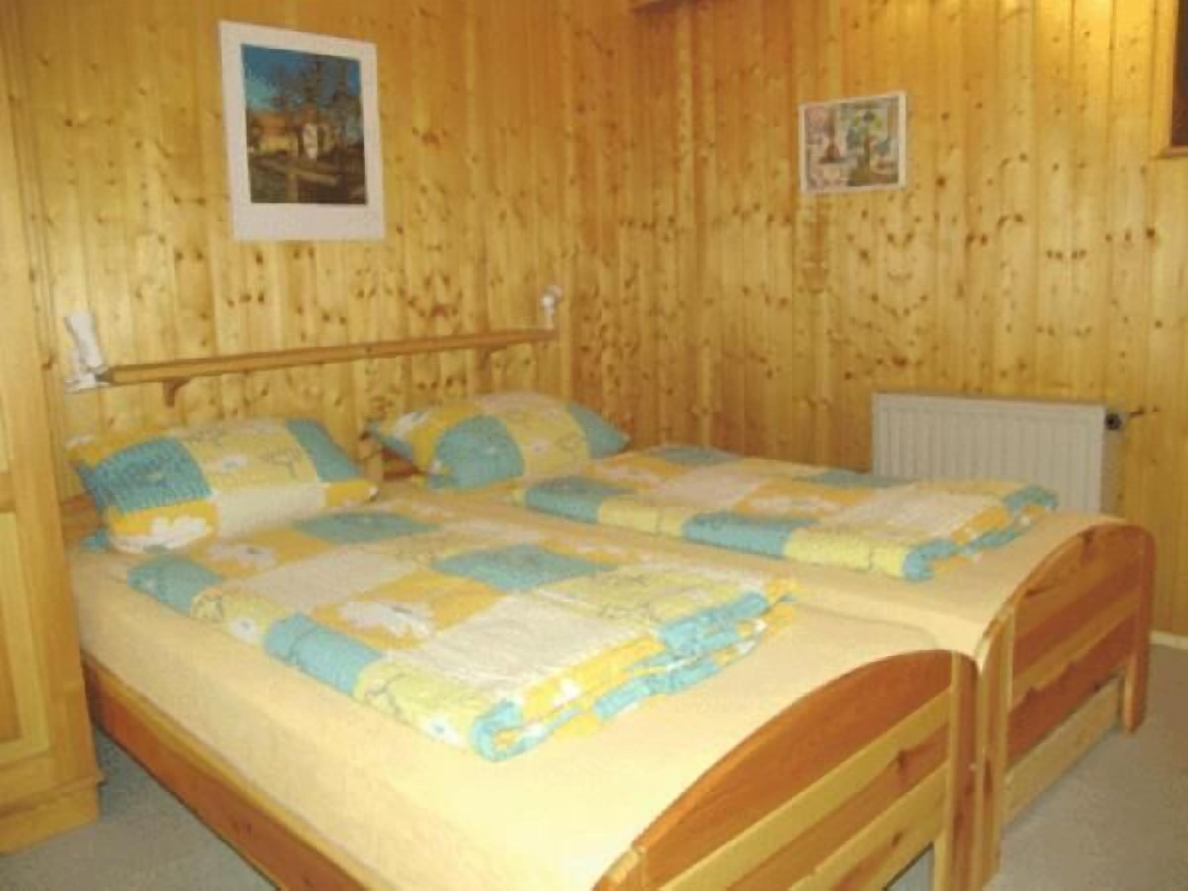A wooden-paneled room with a double bed, two pillows, a nightstand, and a wall-mounted lamp.