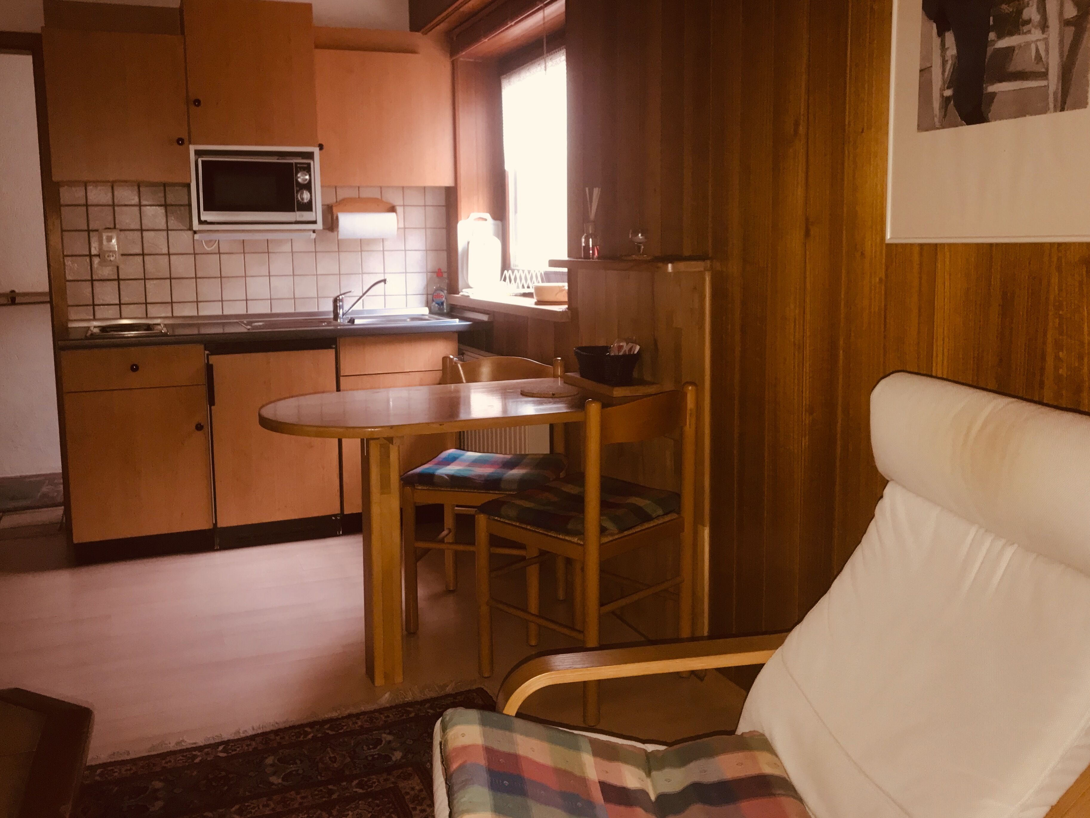 A compact kitchen with a microwave, sink, and wooden cabinets. There is a dining table with a chair and a sofa in the living area.
