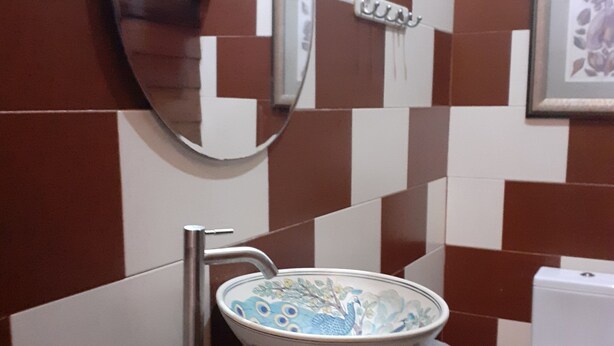 Une salle de bain avec un mur recouvert de carreaux à motifs, un miroir rond, un lavabo agrémenté d’un récipient décoratif, des toilettes et un tabouret en bois.