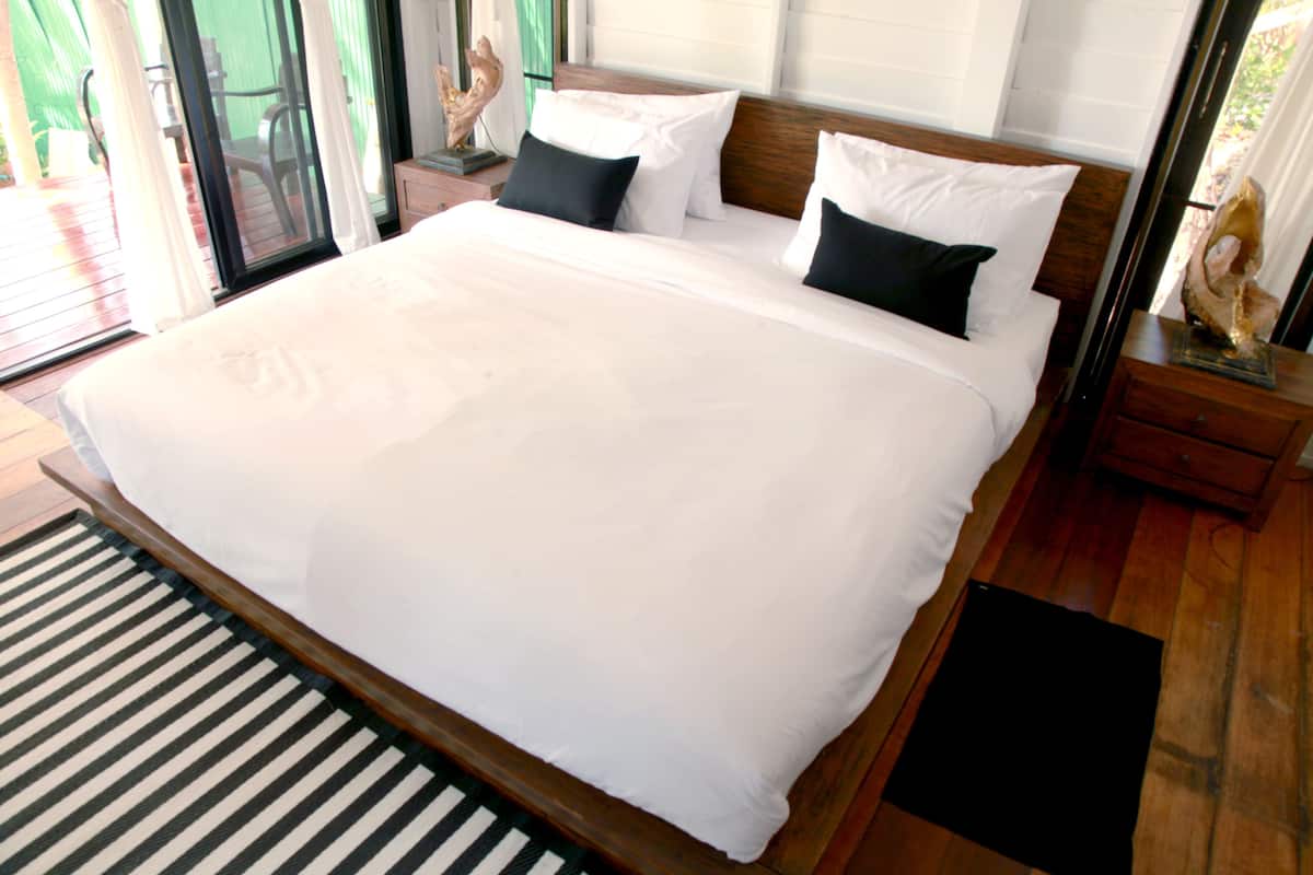 A large bed with white linens and black pillows in a room with wooden flooring and a striped rug.