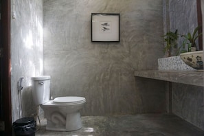A bathroom with a toilet, a framed picture on the wall, and a shelf with a bowl and plants. - Baan Klong Kleng (Ranong)