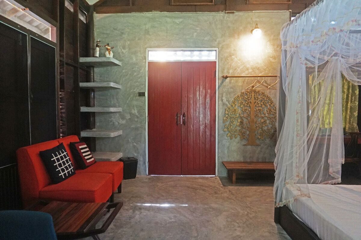 Una habitación con puerta roja, un sofá rojo con cojines estampados, una cama con dosel y un estante montado en la pared con objetos decorativos.