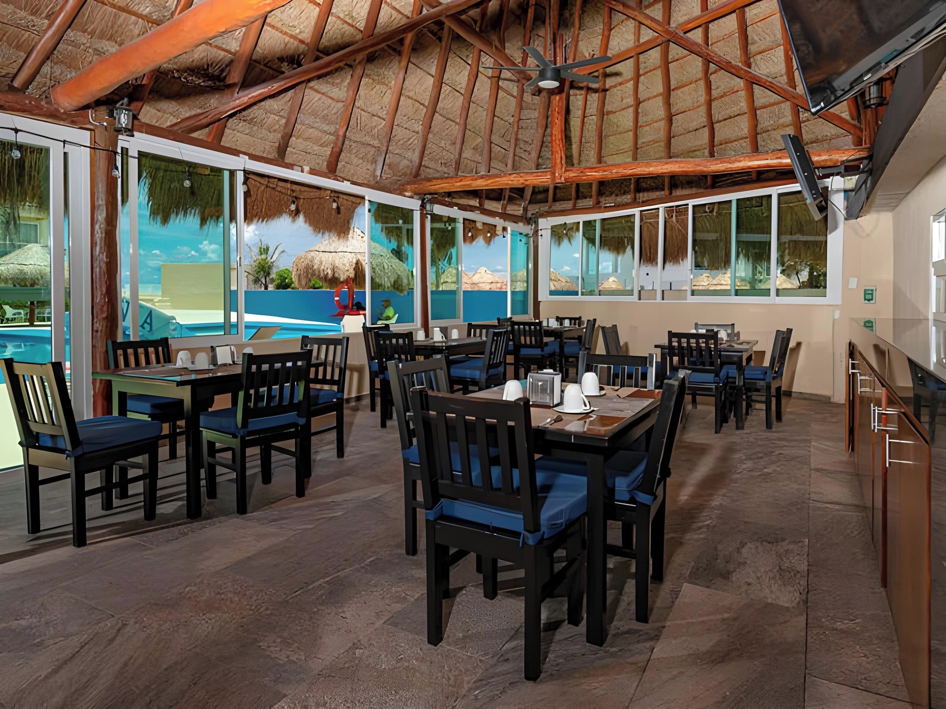 Un comedor interior con mesas y sillas, ventanas grandes y un techo de paja.