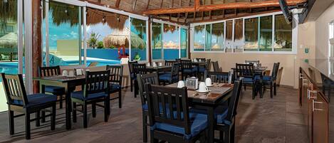 An indoor dining area with tables and chairs, large windows, and a thatched roof.
