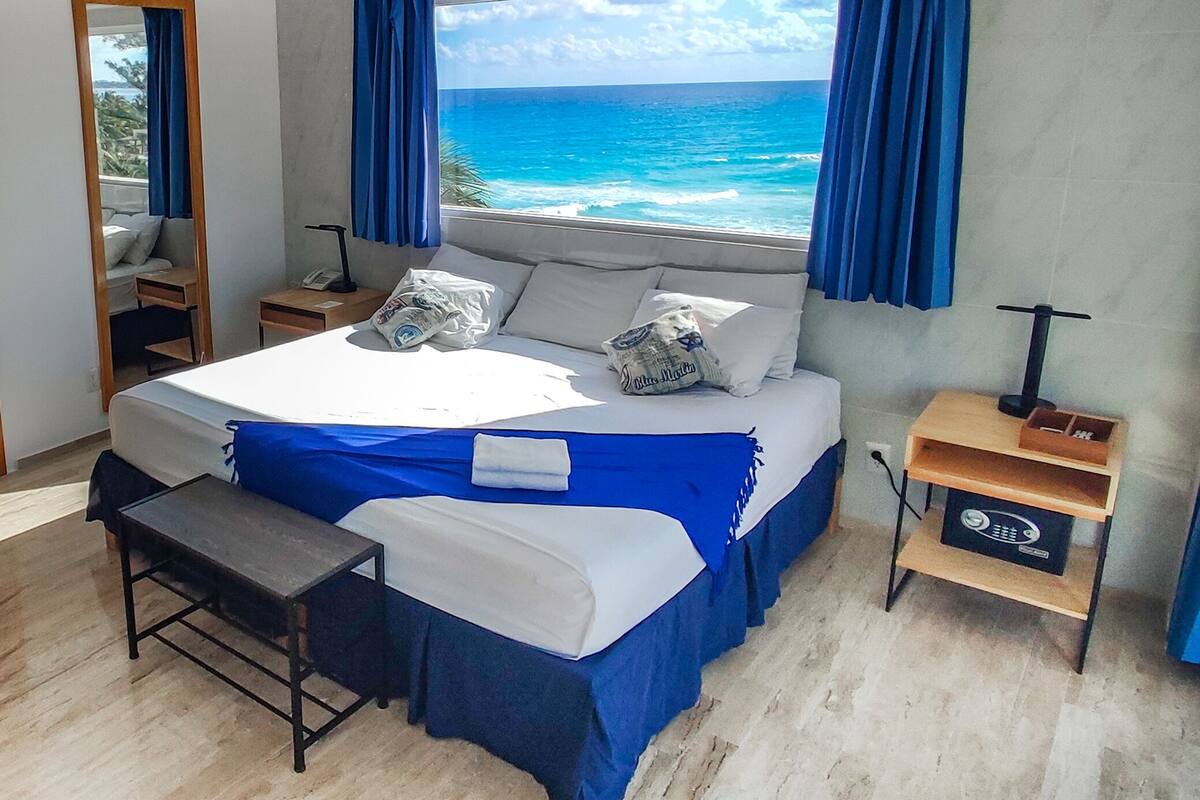 A bedroom with a large bed, a view of the ocean, a small desk, and a nightstand.