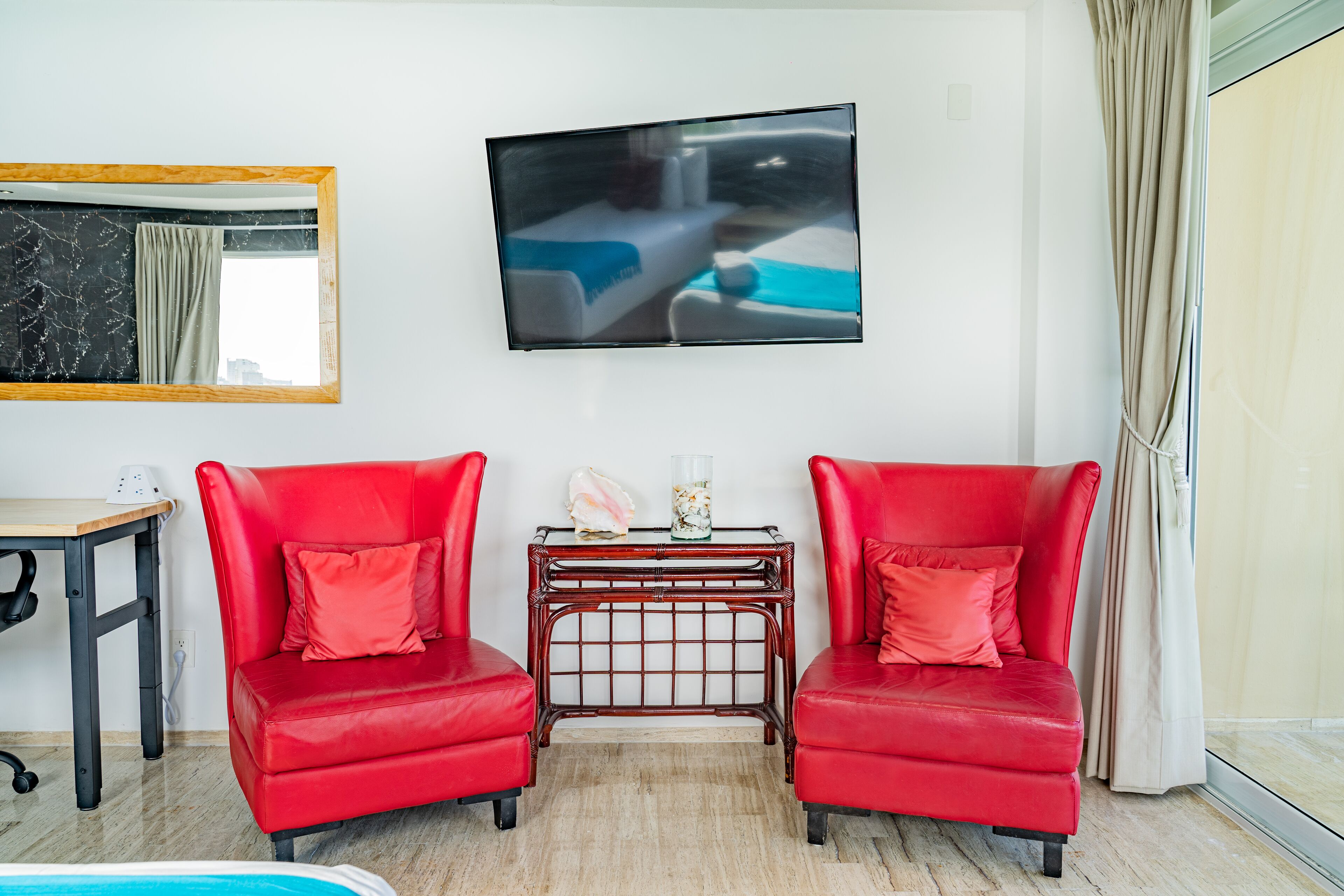 Quarto com duas poltronas de couro vermelho, uma mesa pequena com objetos decorativos, uma televisão de tela plana na parede e uma janela grande com cortinas.