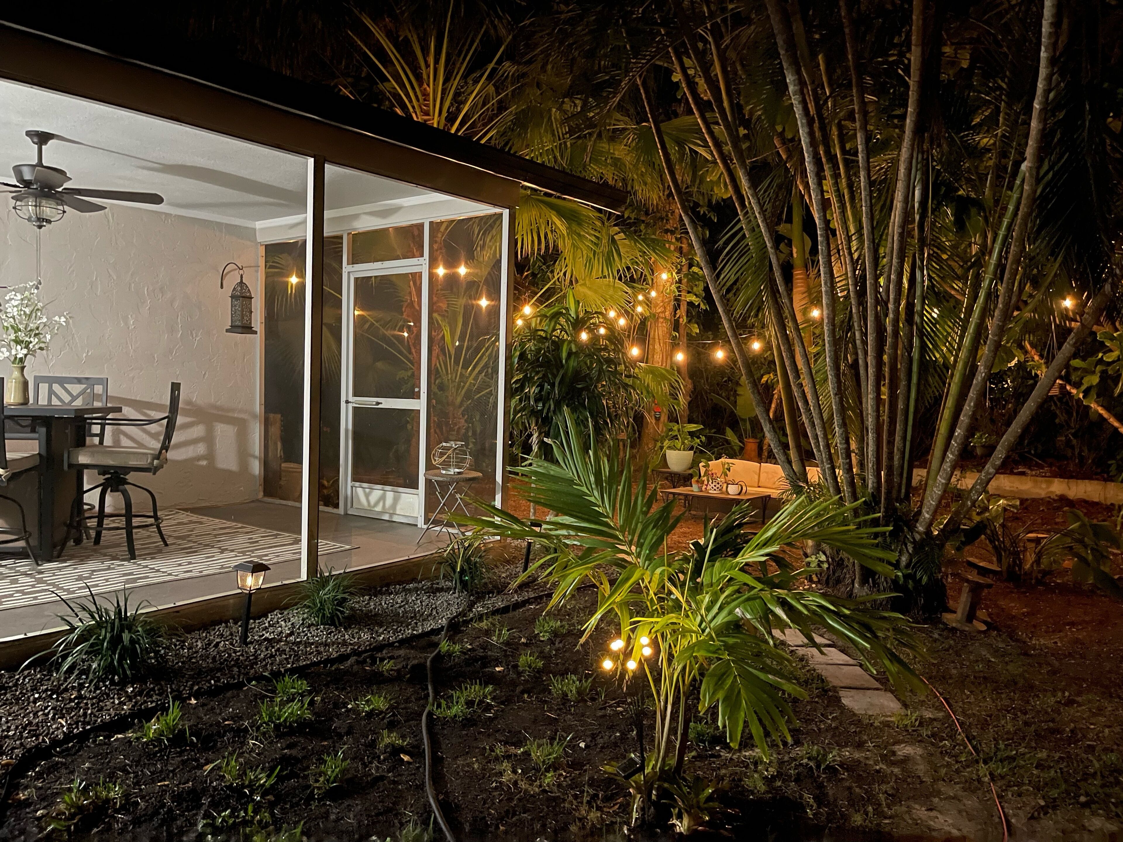 An outdoor patio with a glass door leading to a room, surrounded by lush greenery and string lights.