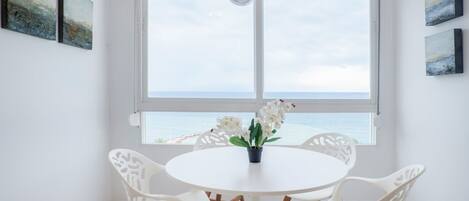 A room with a round white table, white chairs, a potted orchid, and a view of the sea through a large window.