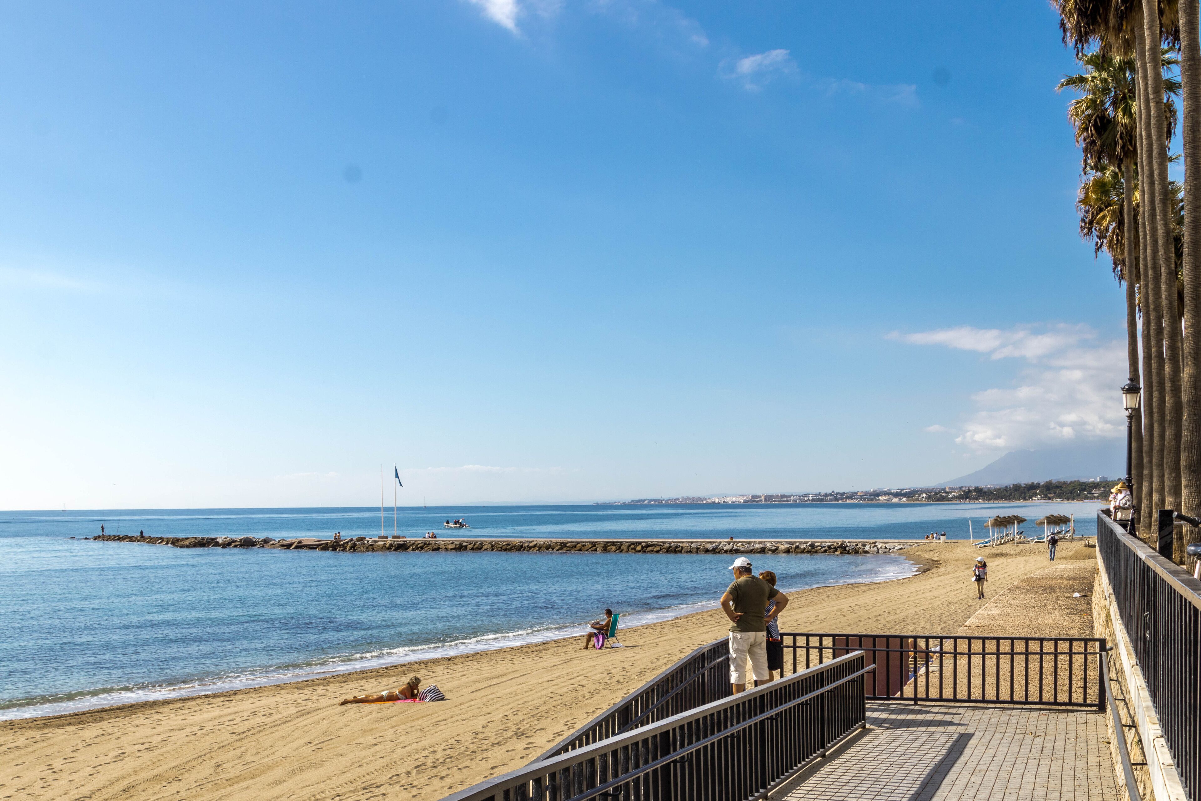 Una playa con un muelle de madera, un rompeolas rocoso y un cielo azul despejado.