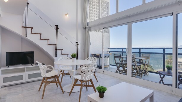 A modern living room with a TV, white furniture, and a balcony with a view of the sea. - Skol Sea Views Apartments (Marbella)
