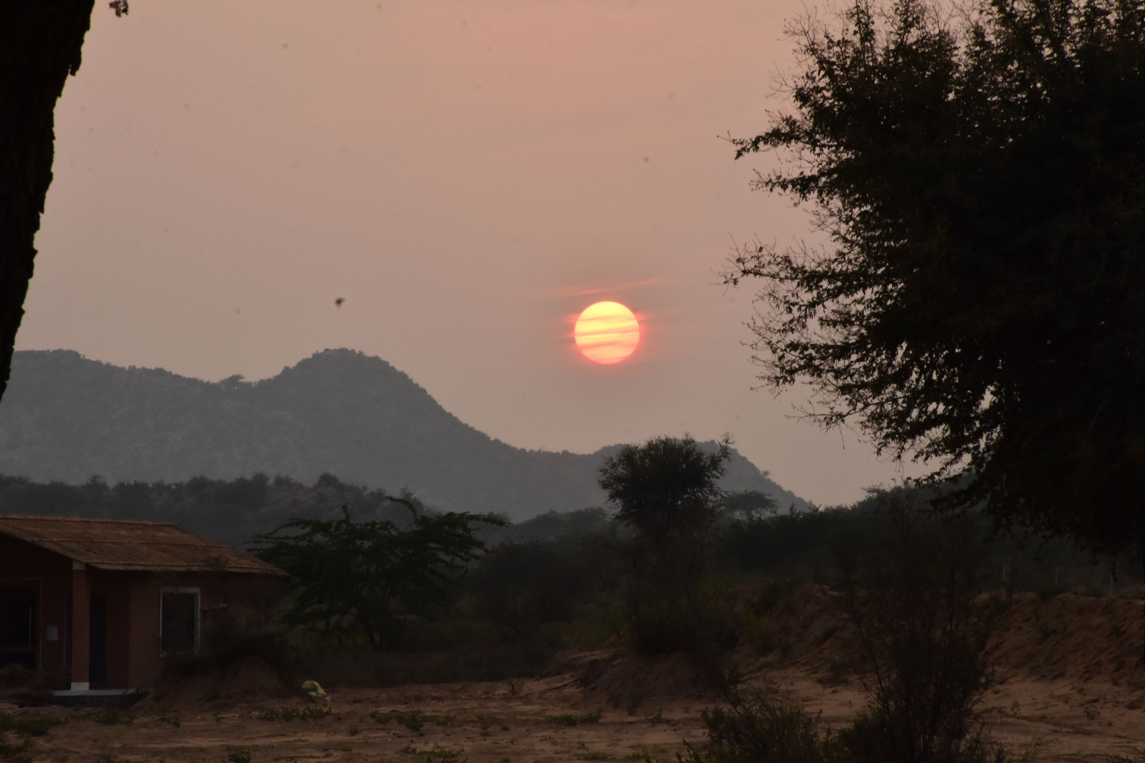 A sunset over a hilly landscape with sparse vegetation and a small building.