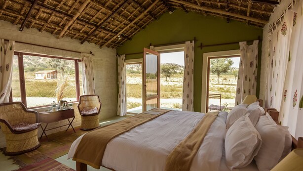 A bedroom with a bed, two wicker chairs, a desk, and a window with curtains. - Camp Land's End (Pushkar)