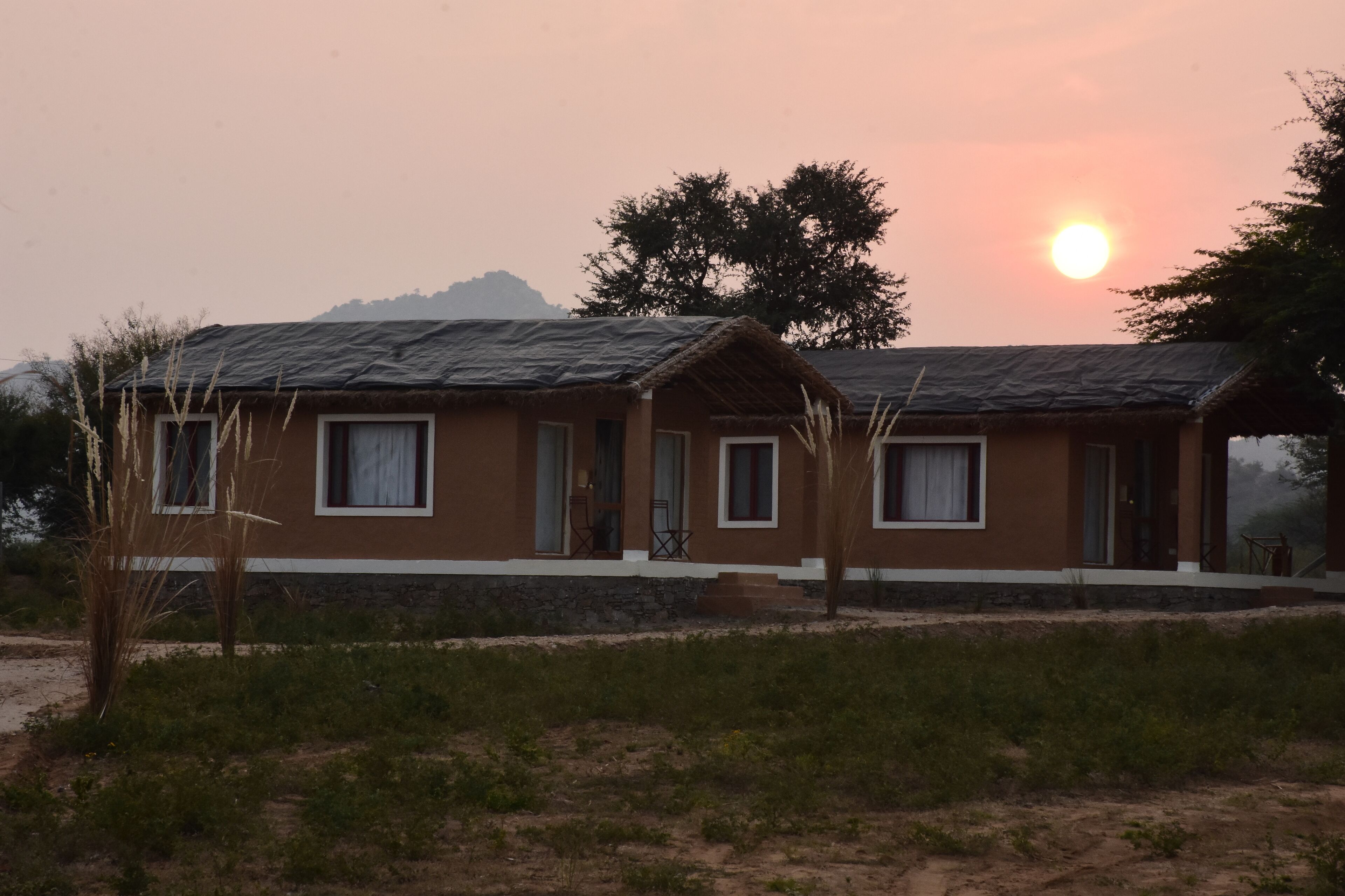 A two-story building with a thatched roof, a small porch, and a view of the sun setting behind a mountain.