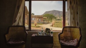 A room with two wicker chairs, a small table with a tea set, and a view of a building and landscape through a window. - Camp Land's End (Pushkar)