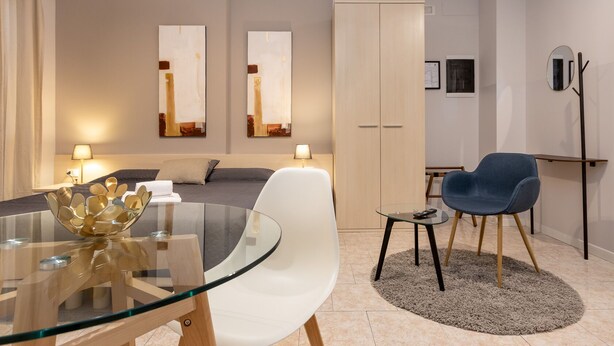 Quarto de hotel moderno com mesa de jantar de vidro, uma cadeira azul, um sofá e um guarda-roupa.
