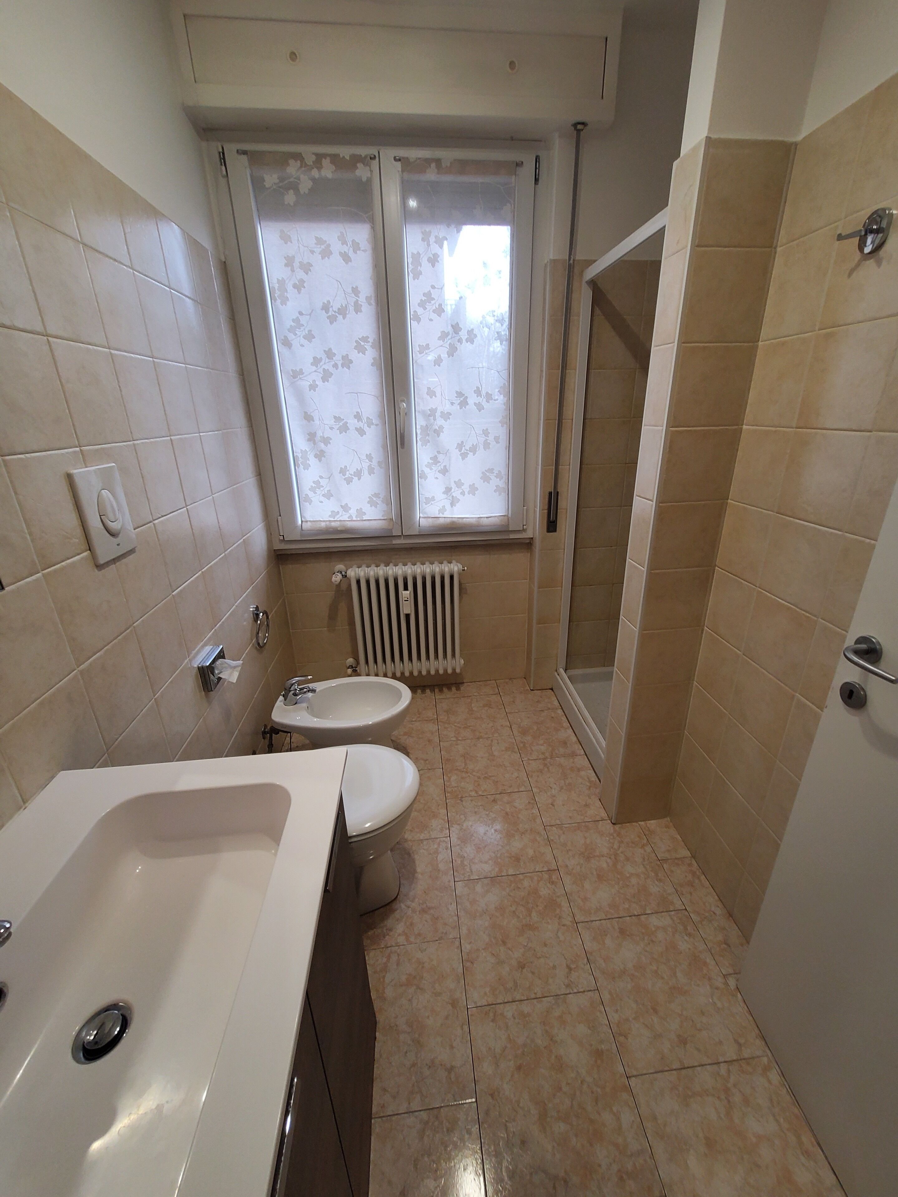 Une salle de bain avec une baignoire, deux toilettes, un radiateur et une fenêtre agrémentée de rideaux à motifs floraux.