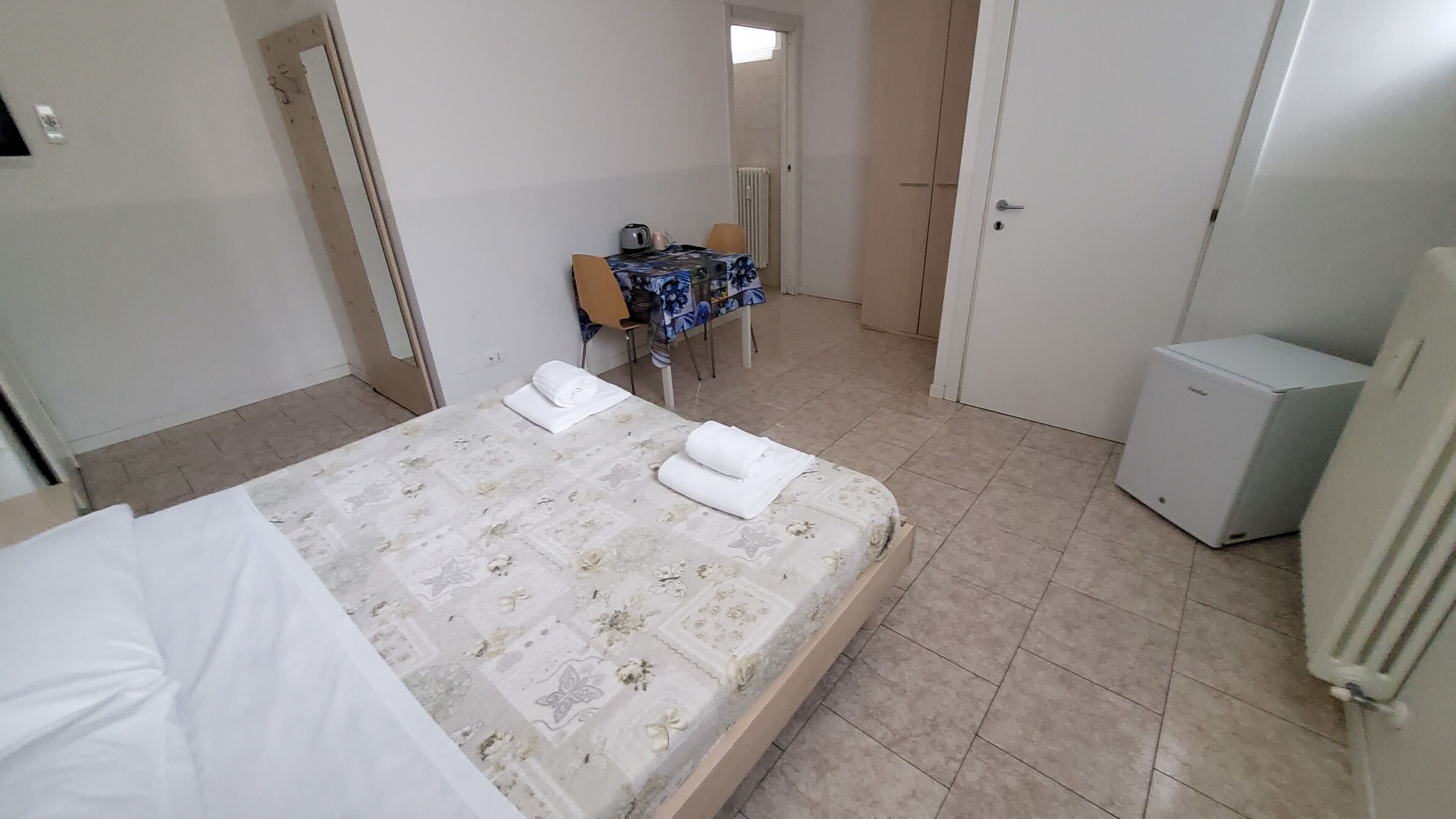 A hotel room with a bed, a small table with chairs, a radiator, and a refrigerator.