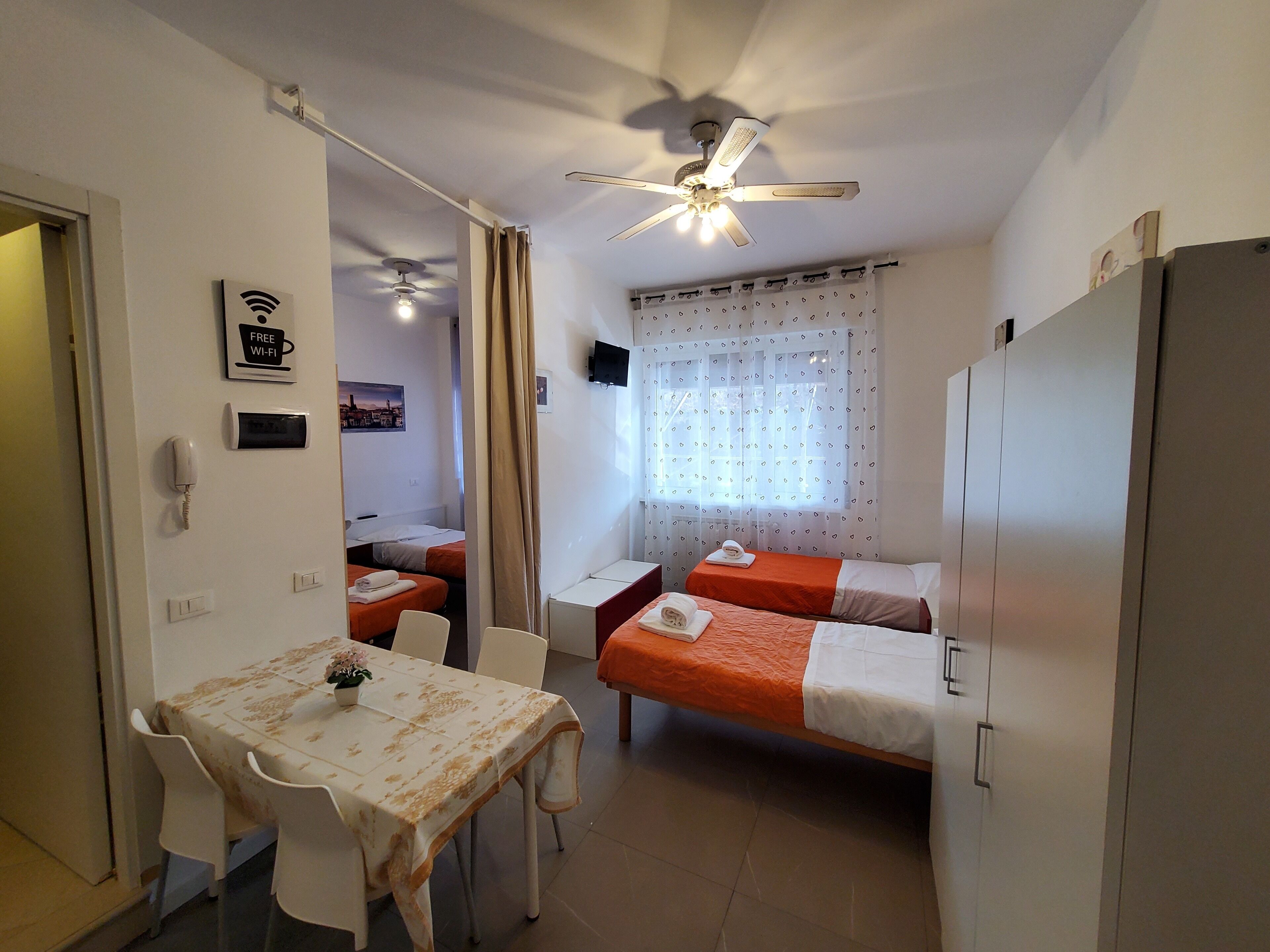 A hotel room with two single beds, a dining table with chairs, a ceiling fan, a TV, and a sign indicating free Wi-Fi.