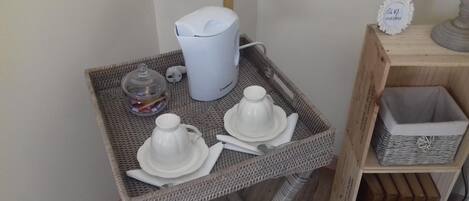 A small wicker table with a white coffee maker, a glass jar, and two white teapots on a tray.